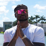 Stylish Ross500  in LosReyes Elevate Bubble gum  sports sunglasses with braided hairstyle and white T-shirt, posing by water with palm trees in background – athletic lifestyle sunglasses for men.