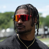Close-up of Ross500 in red elevate cherry LosReyes sunglasses with braided hair and gold chain, looking sideways outdoors – premium sports sunglasses for urban fashion.