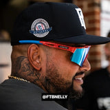 Side-profile of FTB Nelly wearing LosReyes Rocket Pop sunglasses featuring red and blue mirrored shield lenses and wraparound frame, styled with a black fitted Perfect Game baseball cap during tournament weekend