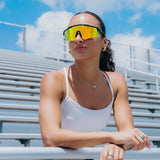 Lifestyle photo of LosReyes Prime Polarized Orange sunglasses worn by female athlete in bleachers, highlighting the vibrant polarized orange mirrored lens and matte black frame designed for baseball, outdoor training, and athletic style.