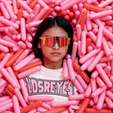Creative lifestyle image of LosReyes Prime Youth Cotton Candy sports sunglasses worn by girl in pink and red capsule pit, emphasizing mirrored UV400 performance lens and playful, youth-focused athletic style