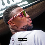 Side-angle lifestyle photo of Saquon Barkley wearing LosReyes pink mirrored shield sunglasses with a wraparound athletic frame, styled during a pro baseball event