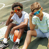 Two kids wearing Candy Collection youth sunglasses with UV400 lenses and colorful candy-themed frames while sitting on a basketball court.