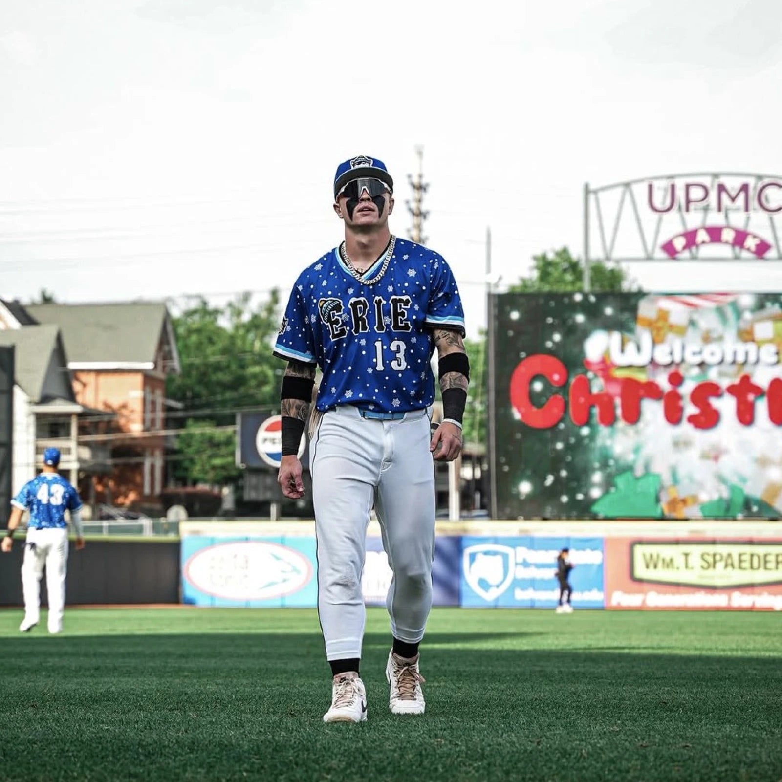 Max Clark standing on the field in an Erie Seawolves holiday uniform, wearing oversized LosReyes SHOWTIME performance shades and a thick chain necklace.