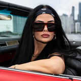 Woman wearing LosReyes Ares omega sunglasses and a headband, sitting in a convertible car with a cityscape background.