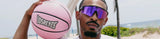 sunglasses man holding a pink LosReyes basketball on the beach, wearing cool uv protection wraparound purple mirrored premium glasses, lifestyle sports eyewear shades