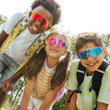 Three children wearing colorful LosReyes sunglasses outdoors with trees in the background
