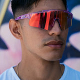 Lifestyle photo of a man wearing LosReyes Prime Galaxy Mercury mirrored sports sunglasses with translucent purple frame and orange shield lens—combining streetwear flair with athletic utility.