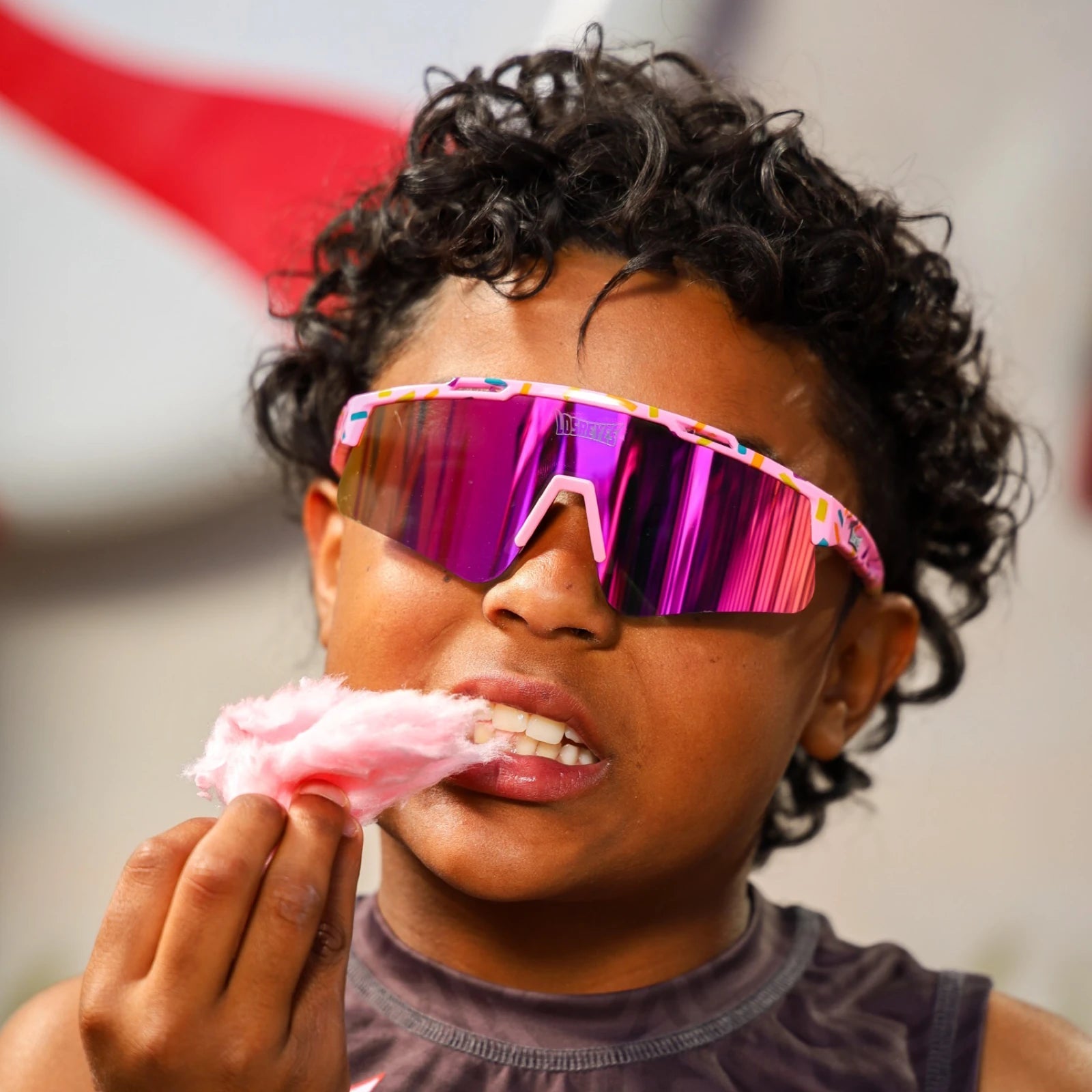 Young boy wearing LosReyes Prime Youth Cream sports sunglasses with pink mirrored UV400 lenses and sprinkle-print frame while eating cotton candy at a summer event