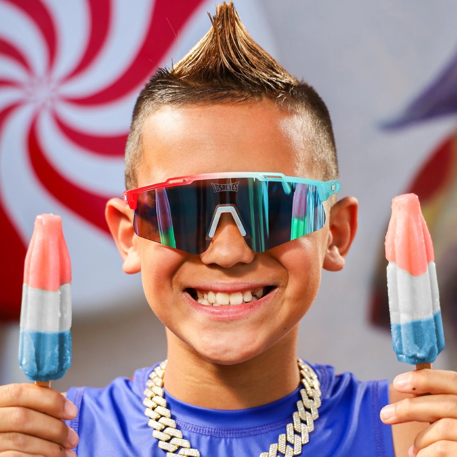 Front view lifestyle of LosReyes Prime Youth Rocket Pop kids sports sunglasses in red, white, and blue with blue mirrored shield lens, modeled by smiling child holding rocket popsicles. UV400 protective eyewear designed for active youth and summer sports style.