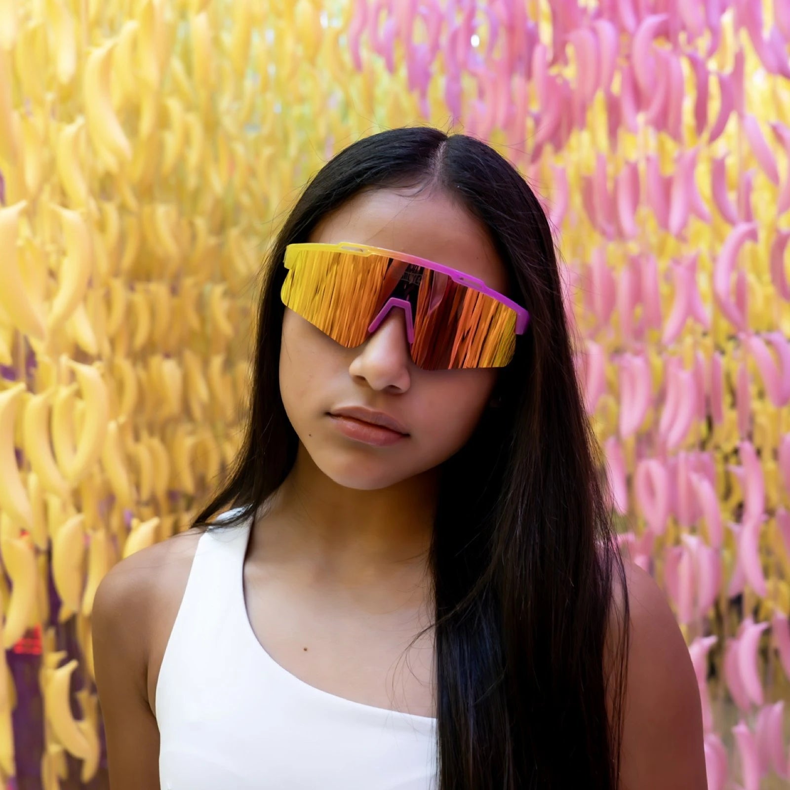 Lifestyle shot of LosReyes Prime Youth Strawberry Banana sports sunglasses with yellow-pink mirrored lens, UV400 shield protection, and bold wraparound frame — styled in banana-themed art room.