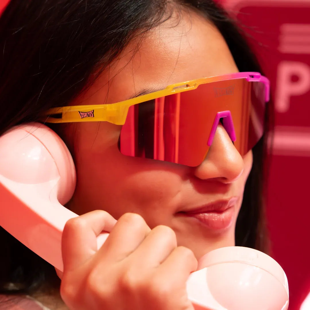 Close-up lifestyle photo of LosReyes Prime Youth Strawberry Banana sports sunglasses featuring pink and yellow mirrored shield lens, wraparound performance frame, and UV400 protection — worn by girl holding retro phone.