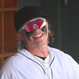 Max Clark smiling in the dugout wearing LosReyes sunglasses with pink reflective lenses and teal frame, dressed in a white jersey with eye black makeup.
